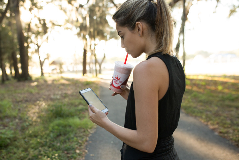 A woman pausing after a jog and drinking Smoothie King while looking at her phone