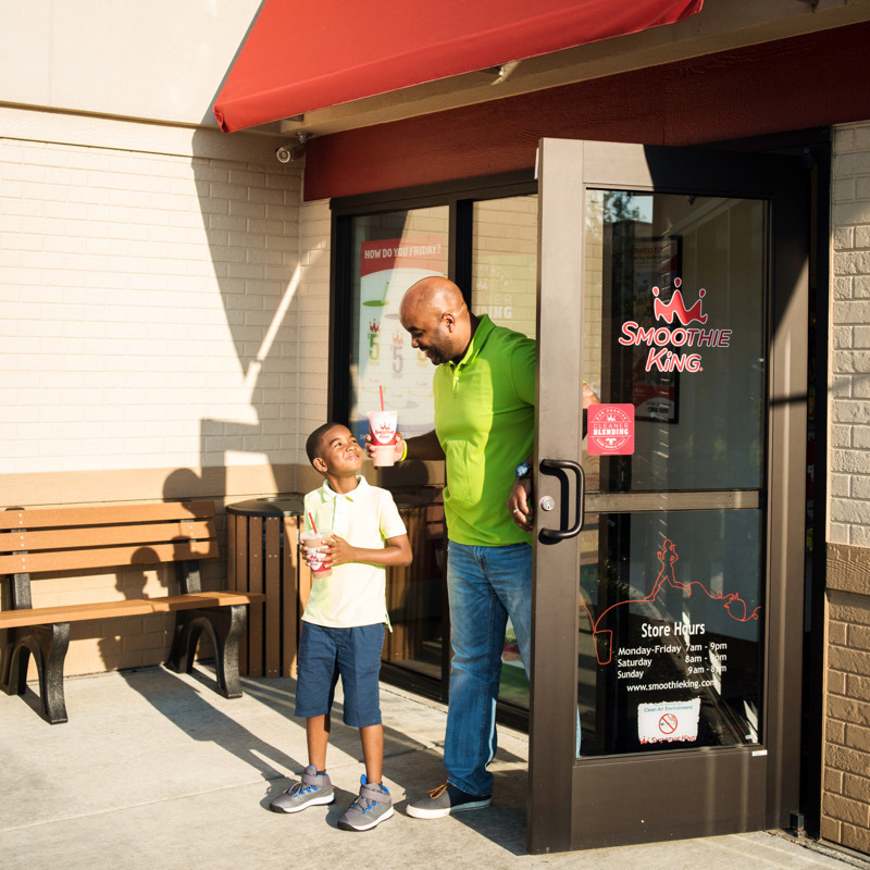 A father and son exit a Smoothie King store with their smoothies in hand