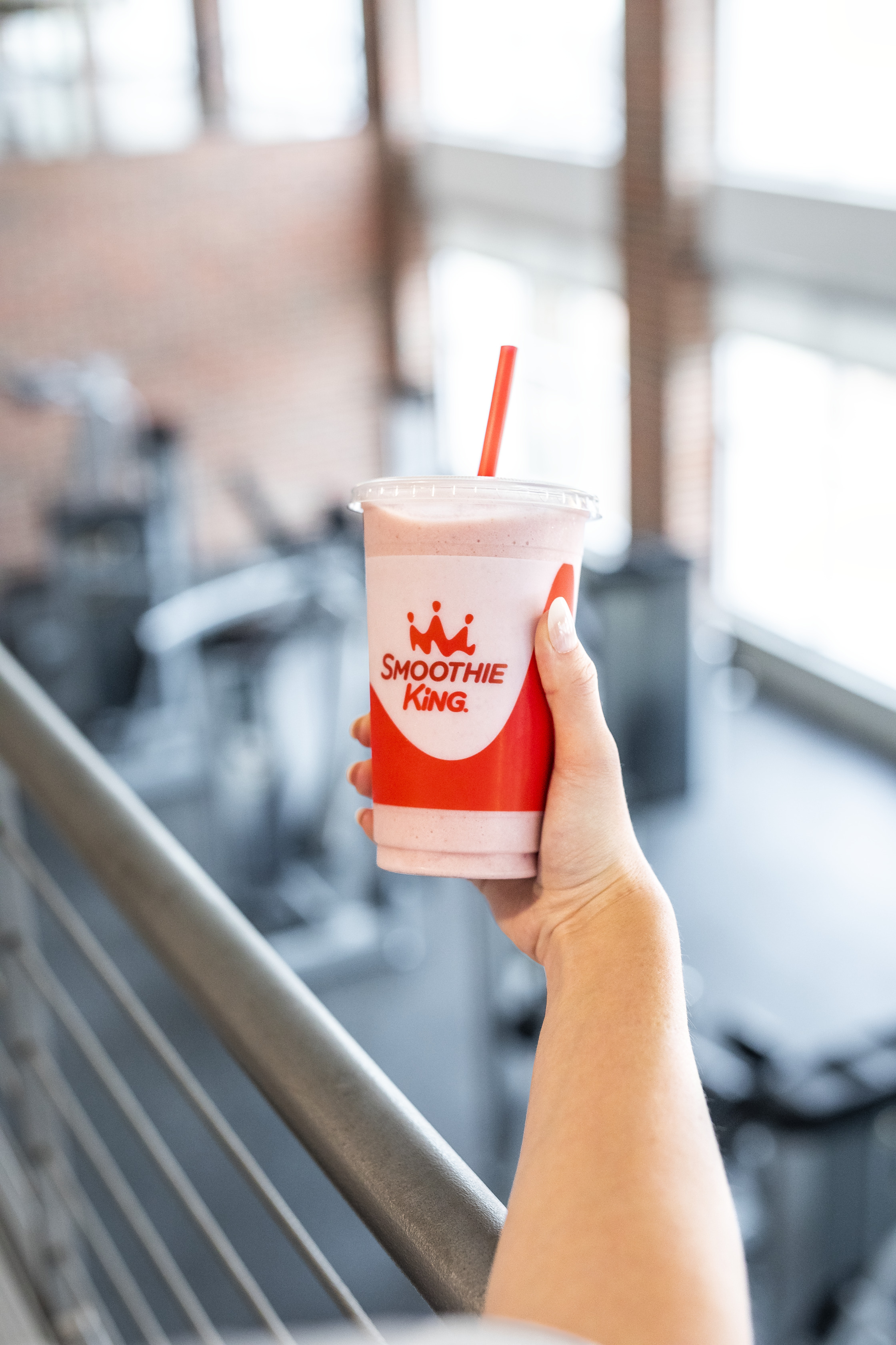 A woman holding a strawberry smoothie from Smoothie King with her outstretched arm over a railing at a gym