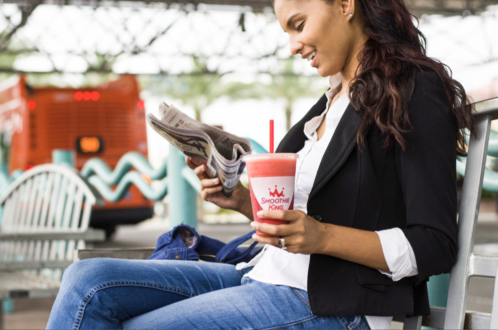 A woman reading a paper smiles while she sits on a bench and drinks an energy smoothie with multivitamins