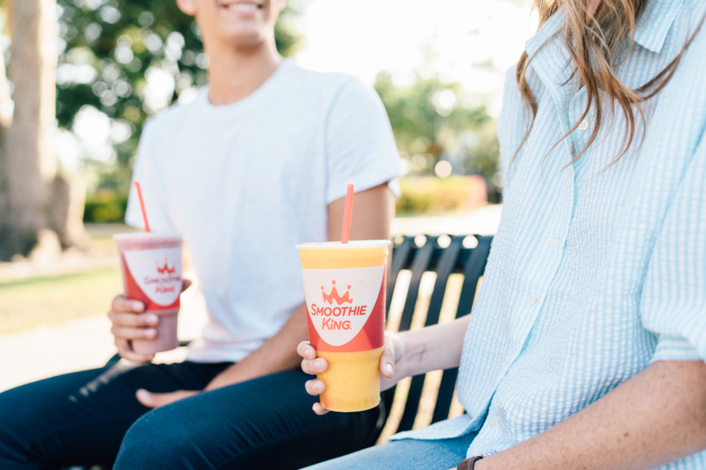 A smiling couple drink Smoothie King smoothies at a park while sitting on a bench