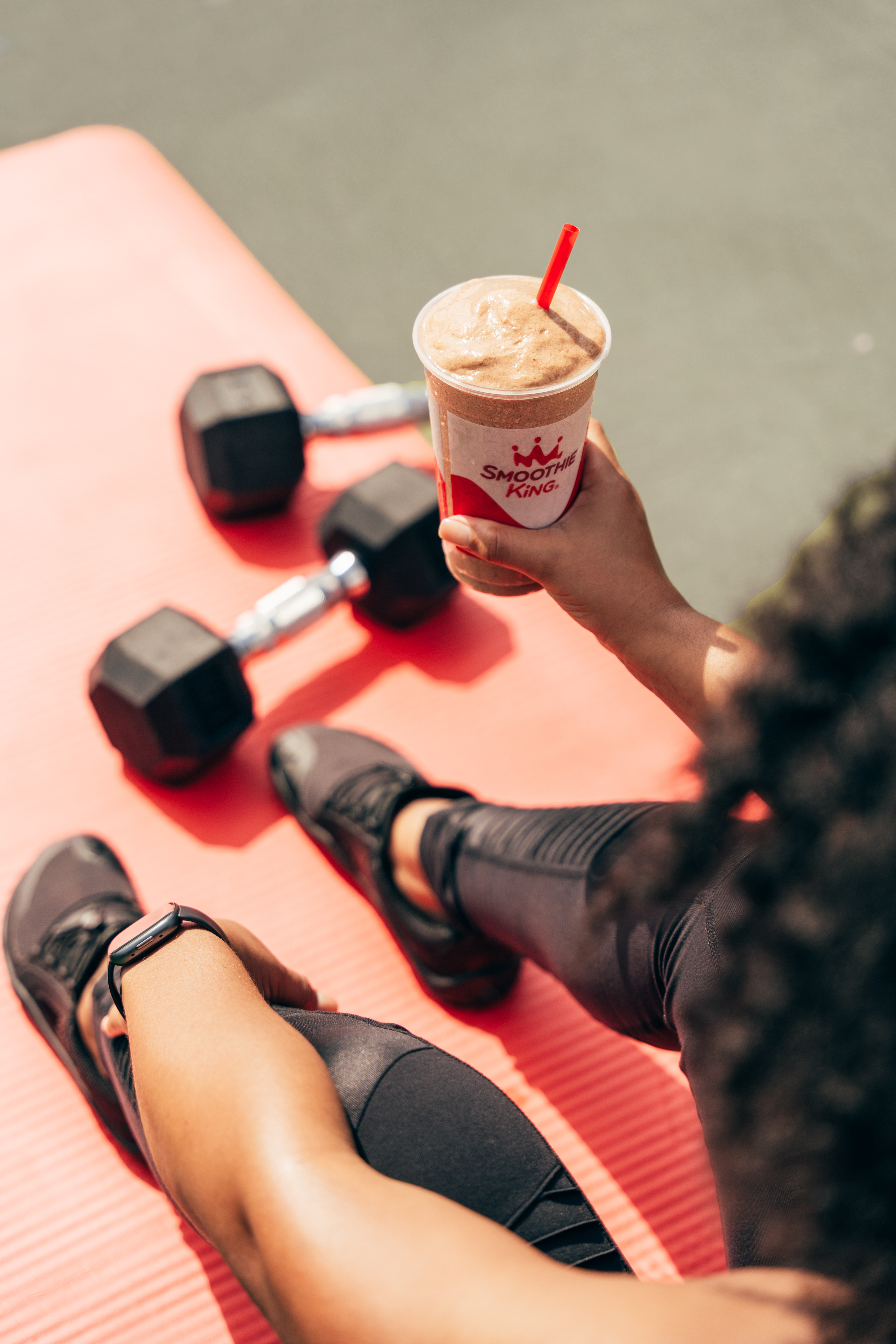 A woman enjoying her post-workout chocolate smoothie from Smoothie King after working out with dumbbells on a mat